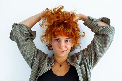 portrait red-haired curly attractive teenage girl with emotions on face, holding hair with hands, wearing plaid men's shirt, against wall