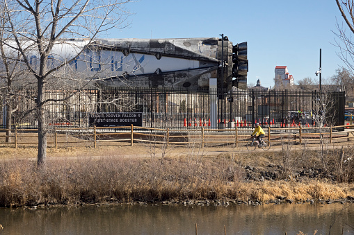 Riding his bike along the South Platte River Trail, a man passes by a huge and charred Falcon 9 SpaceX rocket booster lying on its side along the river near downtown Littleton Colorado. The courthouse and grain elevator rise in the distance.