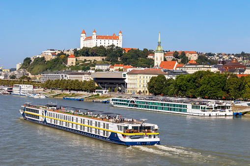 River cruise ship arriving on Danube in Bratislava with old town cityscape in background and other ships docked along embankment