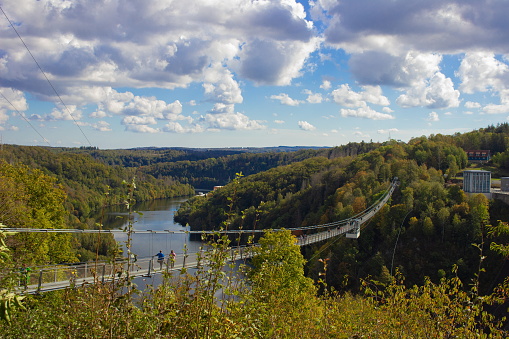 The suspension bridge connects two forested hillsides with autumn foliage under a clear sky, with people walking across while admiring the river below. The lush trees in the valley display a variety of fall colors, and a small building is visible on the right. The bridges cables create a delicate silhouette against the bright sky.