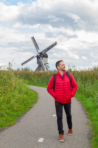 Happiness young latin male tourist taking a euro trip around Windmills in Kinderdijk, South Holland, Netherlands.\n\nA breathtaking beautiful inspirational landscape with a collection of 18th Century traditional Dutch windmills at afternoon. Fascinating places, tourist attraction.\n\nHe's wearing a red sweater, black jeans and carry a black backpack over his shoulders.