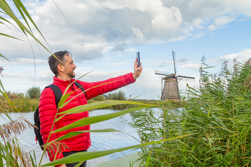 Happiness young latin male tourist taking a euro trip around Windmills in Kinderdijk, South Holland, Netherlands. He's taking a selfie with his smart phone using 5G technology.

A breathtaking beautiful inspirational landscape with a collection of 18th Century traditional Dutch windmills at afternoon. Fascinating places, tourist attraction.

He's wearing a red sweater, black jeans and carry a black backpack over his shoulders.