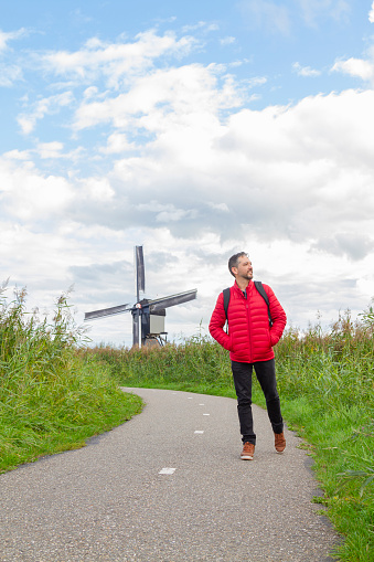 Happiness young latin male tourist taking a euro trip around Windmills in Kinderdijk, South Holland, Netherlands.

A breathtaking beautiful inspirational landscape with a collection of 18th Century traditional Dutch windmills at afternoon. Fascinating places, tourist attraction.

He's wearing a red sweater, black jeans and carry a black backpack over his shoulders.