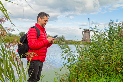 Happiness young latin male tourist taking a euro trip around Windmills in Kinderdijk, South Holland, Netherlands. He's browsing the net looking for information on his smart phone using 5G technology.

A breathtaking beautiful inspirational landscape with a collection of 18th Century traditional Dutch windmills at afternoon. Fascinating places, tourist attraction.

He's wearing a red sweater, black jeans and carry a black backpack over his shoulders.