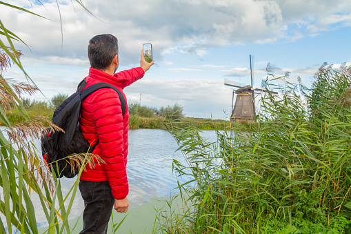 Happiness young latin male tourist taking a euro trip around Windmills in Kinderdijk, South Holland, Netherlands. He's taking a selfie with his smart phone using 5G technology.

A breathtaking beautiful inspirational landscape with a collection of 18th Century traditional Dutch windmills at afternoon. Fascinating places, tourist attraction.

He's wearing a red sweater, black jeans and carry a black backpack over his shoulders.