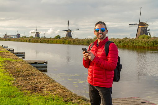 Happiness young latin male tourist taking a euro trip around Windmills in Kinderdijk, South Holland, Netherlands. He's browsing the net looking for information on his smart phone using 5G technology.

A breathtaking beautiful inspirational landscape with a collection of 18th Century traditional Dutch windmills at afternoon. Fascinating places, tourist attraction.

He's wearing a red sweater, black jeans and carry a black backpack over his shoulders.