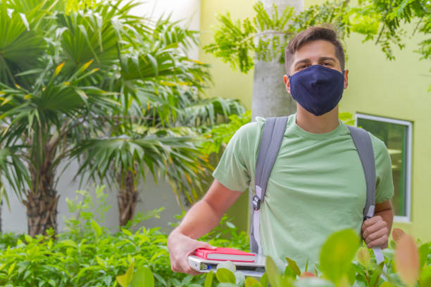 portrait of young latin college happy handsome university male student carrying a backpack and book walking in university campus outside the library, wearing a green t-shirt and blue pants. - library student latin american and hispanic ethnicity university stock-fotos und bilder