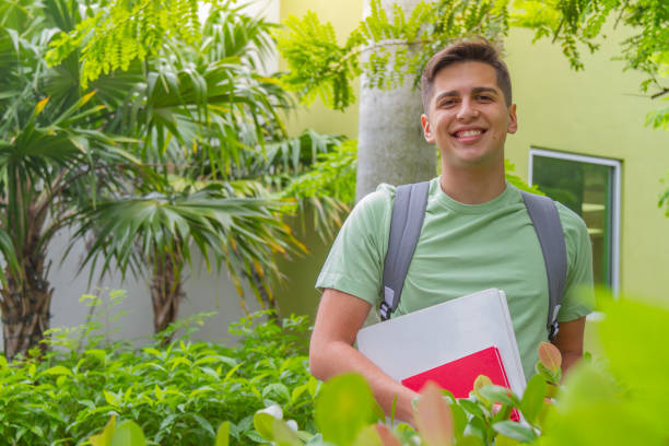 portrait of young latin college happy handsome university male student carrying a backpack and book walking in university campus outside the library, wearing a green t-shirt and blue pants. - library student latin american and hispanic ethnicity university stock-fotos und bilder