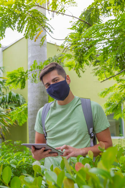 portrait of young latin college happy handsome university male student carrying a backpack and holding a digital tablet while surfing the net with 5g technology in university campus outside the library. - library student latin american and hispanic ethnicity university stock-fotos und bilder