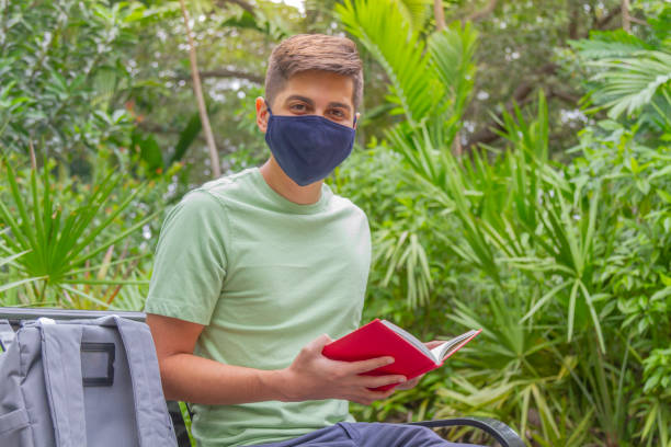 portrait of young latin college happy handsome university male student carrying a backpack and reading a book in university campus outside the library. - library student latin american and hispanic ethnicity university stock-fotos und bilder