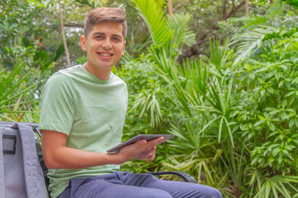 porträt eines jungen, glücklichen, gutaussehenden studenten, der einen rucksack und ein buch trägt und auf dem universitätscampus vor der bibliothek spazieren geht, in einem grünen t-shirt und einer blauen hose. - library student latin american and hispanic ethnicity university stock-fotos und bilder