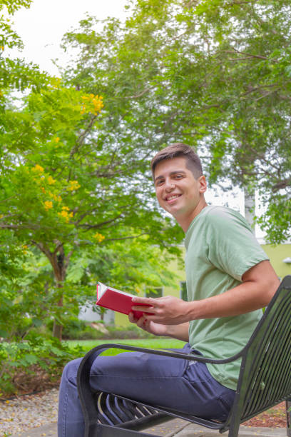 portrait of young latin college happy handsome university male student carrying a backpack and reading a book in university campus outside the library. - library student latin american and hispanic ethnicity university stock-fotos und bilder