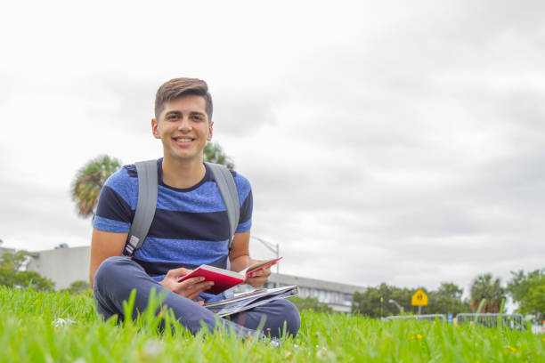 portrait of young latin college happy handsome university male student carrying a backpack and reading a book over the grass in university campus outside the library, wearing a blue t-shirt and pants. - library student latin american and hispanic ethnicity university stock-fotos und bilder