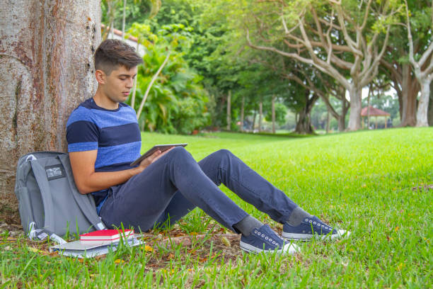 porträt eines jungen, gutaussehenden studenten, der einen rucksack trägt und ein digitales tablet in der hand hält, während er auf dem universitätscampus vor der bibliothek mit 5g-technologie im internet surft und ein blaues t-shirt und eine blaue hos - library student latin american and hispanic ethnicity university stock-fotos und bilder