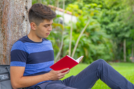 Portrait of young latin college happy handsome university male student carrying a backpack and reading a book over the grass in university campus outside the library, wearing a blue t-shirt and pants.\n\nEducation concept.  Copy Space.