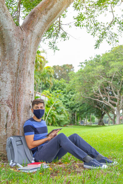 portrait of young latin college happy handsome university male student carrying a backpack and holding a digital tablet while surfing the net with 5g technology in university campus outside the library. - library student latin american and hispanic ethnicity university stock-fotos und bilder