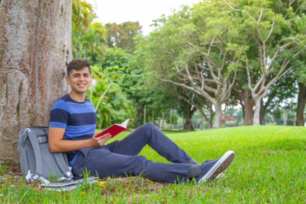 portrait of young latin college happy handsome university male student carrying a backpack and reading a book over the grass in university campus outside the library, wearing a blue t-shirt and pants. - library student latin american and hispanic ethnicity university stock-fotos und bilder