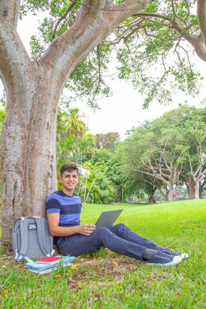 portrait of young latin college happy handsome university male student carrying a backpack and holding a laptop while surfing the net with 5g technology, sitting over the grass in university campus outside the library, wearing a blue t-shirt and pants. - library student latin american and hispanic ethnicity university stock-fotos und bilder