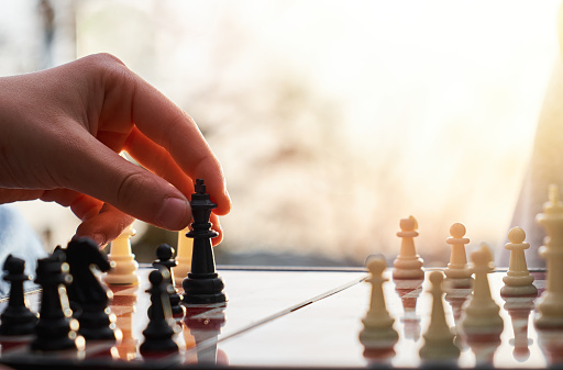 The chess board is made of wood. Person playing chess in nature. The person is holding a black king in their hand.