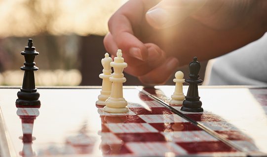 The chess board is made of wood. Person playing chess in nature. The person is holding a black king in their hand.