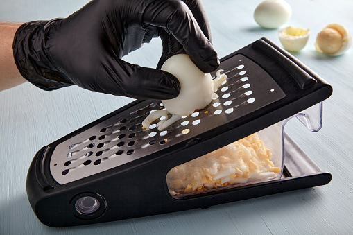Egg chopped on a grater. Hands in black latex gloves rub a hard boiled chicken egg.