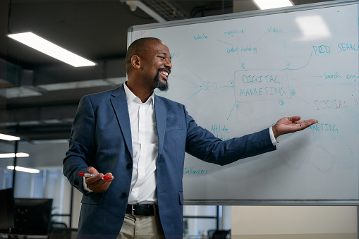 Happy mature black businessman smiling and gesturing next to whiteboard during meeting in corporate office