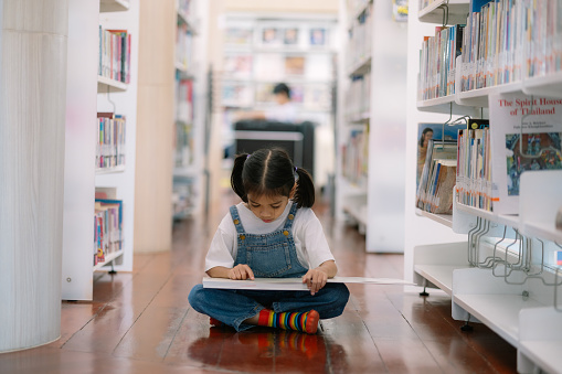 A young girl is sitting on the floor in a library, reading a book. Concept of quiet and concentration, as the girl is focused on her reading. The library setting suggests a peaceful