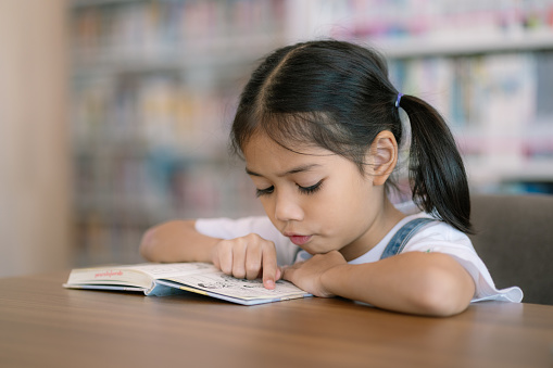A young girl is reading a book at a table. She is looking at the page and pointing at something. The scene is set in a library, with many books visible in the background