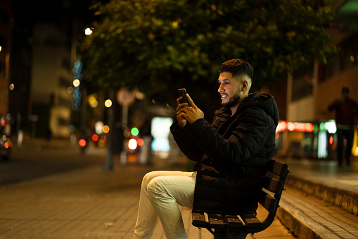 Young man leaves work and sits on the street to check some emails and messages on his cell phone and answer messages to his family and friends before leaving on his way home