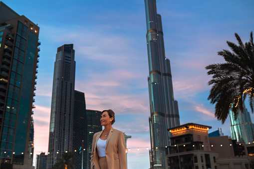 Asian businesswoman wearing beige jacket on business trip exploring city at night, smiling and looking up admiring building architecture in city center, standing against background of modern skyscrapers