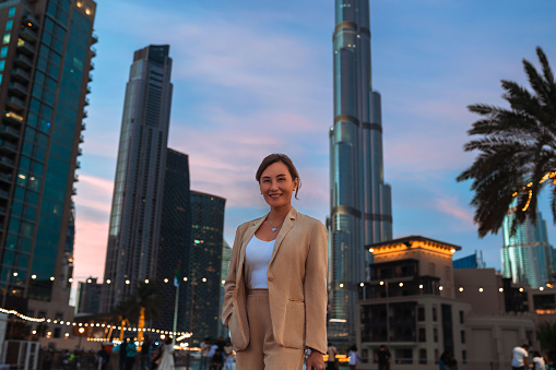 Asian businesswoman wearing beige jacket and pants on business trip, smiling cheerfully at camera, standing against background of modern skyscrapers in city center at night