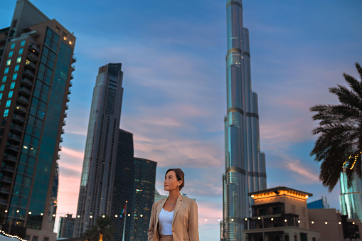 Asian businesswoman wearing beige jacket over white top, standing outdoors looking up at ultramodern skyscrapers, admiring building architecture in city center at night