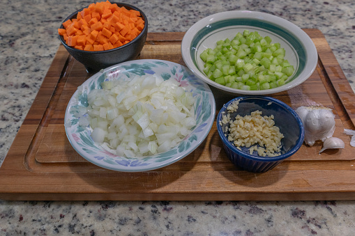 side view of chopped carrots, celery, onions and garlic in bowls with whole garlic on cutting board with copy space