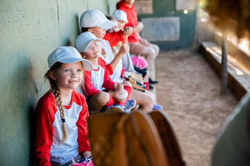 A dugout scene captures six elementary school-aged children, outfitted in baseball team uniforms, seated in anticipation