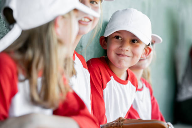 A candid shot captures six elementary school children, dressed in baseball team uniforms, enjoying a moment of camaraderie in the dugout