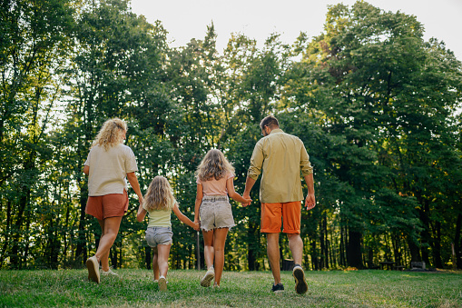 Back view of an adventurous family of four holding hands and exploring nature together. A young explorer family is hiking in nature. Rear view of family of four holding hands and exploring nature.