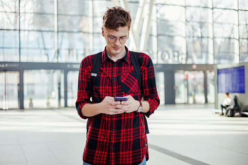 Portrait of a hipster traveler standing at metro station with a backpack and typing messages on his phone. An urban, trendy passenger is texting sms at subway.A commuter using phone for communication.