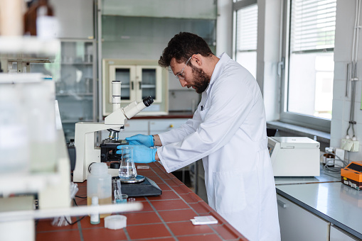 A young male Hispanic scientist wearing a white lab coat, protective gloves and eyewear is working with a microscope at a laboratory. He is working focused at a lab full with lab equipment.