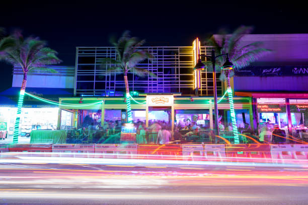 Night view of light trails from passing cars on Collins Avenue in Miami Beach with palm trees and buildings in the background.