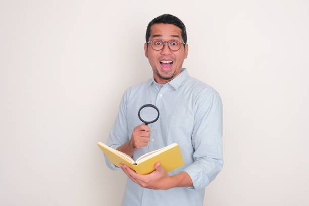 istock Adult Asian man showing wow face expression while holding a book and magnifying glass 2128858424
