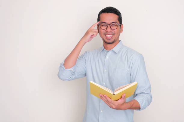 istock Adult Asian man smiling and pointing his temple while holding a book 2128882626