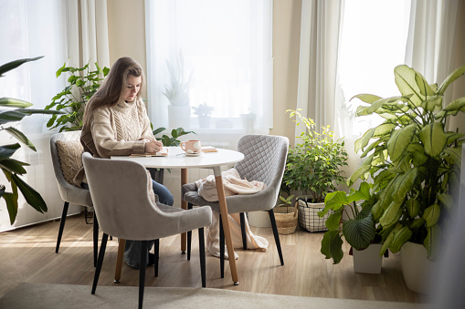 Dreaming plus size woman relaxing at comfortable room with houseplant writing notes in diary sitting at table. Smiling domestic female planning work agenda schedule appointment at cozy home window