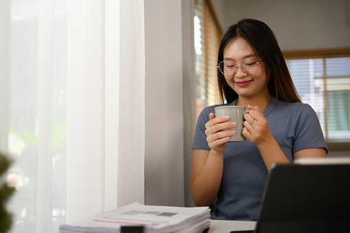 Happy young Asian female freelancer enjoying her cup of coffee while working at home office.