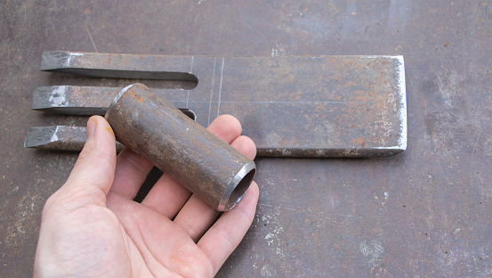 A hand holding an iron pipe with two short metal nails on it, next to the flat rectangular piece of steel lying in front of them, which is used for making scrap and branch cutters.