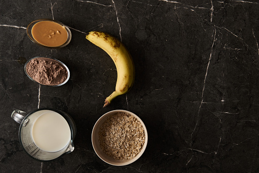 Ingredients for a Protein-Rich Smoothie: Fresh Banana, Rolled Oats, Creamy Milk, and Chocolate Protein Powder, Artistically Placed on a Luxurious Black Marble Surface. High quality photo