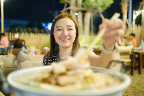 Happy delightful Asian woman enjoy eating the Thai Barbecue or Moo Kata buffet. Woman using chopstick picking up a piece of sliced belly pork from the BBQ pan. Tradition - modern Thai food concept.