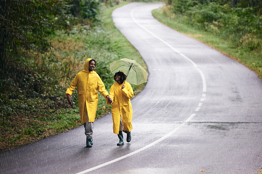 Carefree African American couple holding hands while walking on the road during rainy autumn day. Copy space.
