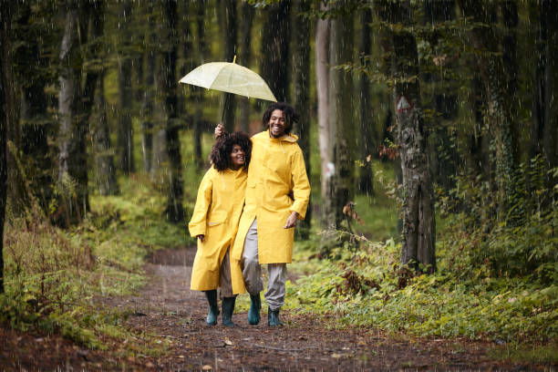 happy black couple in raincoats walking on rain through the woods. - candid women african descent umbrella foto e immagini stock