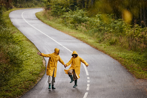 Carefree black couple in raincoats holding hands and having fun during their walk on rainy autumn day on the road. Copy space.