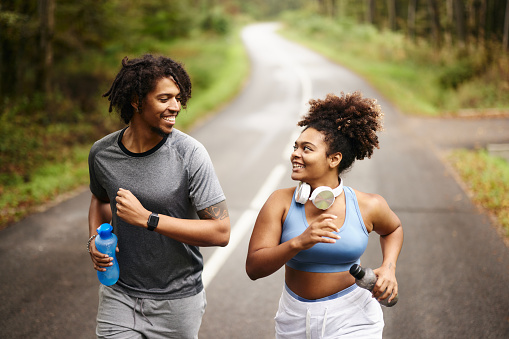 Happy African American athletic couple talking while jogging in autumn day on empty road.